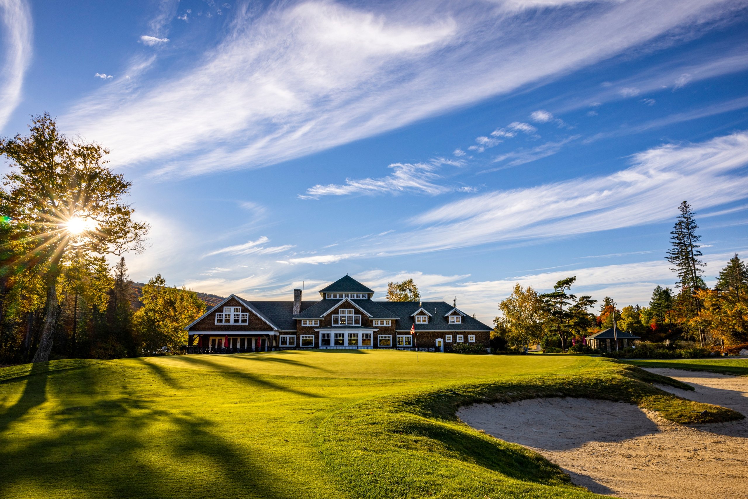A large lodge-style building sits on a hilltop of lush green grass with the sun shining through the trees at Mount Washington Resort and Spa.