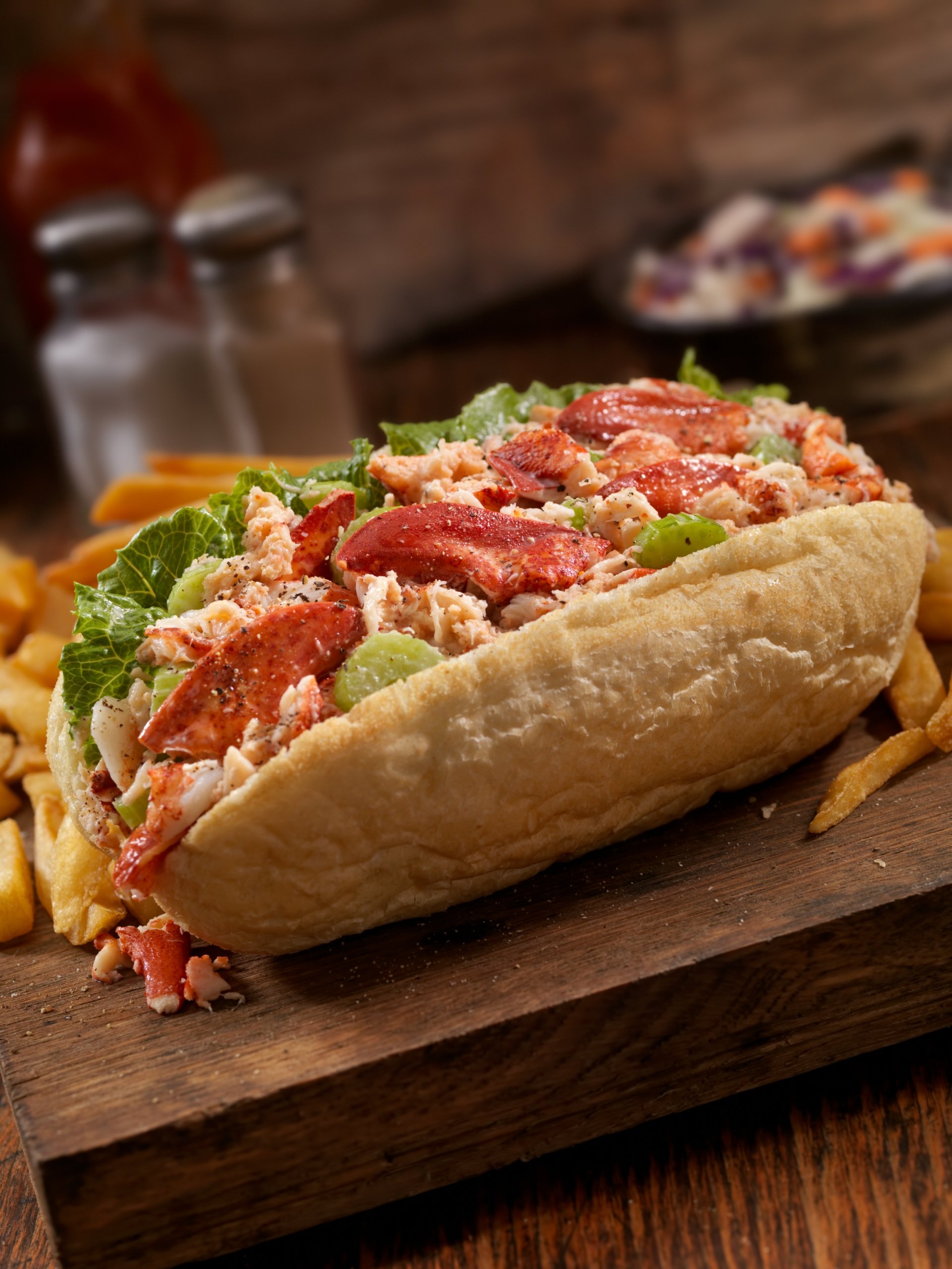 Close-up of a lobster roll filled with fresh lobster meat and lettuce, served with a side of fries on a wooden board.