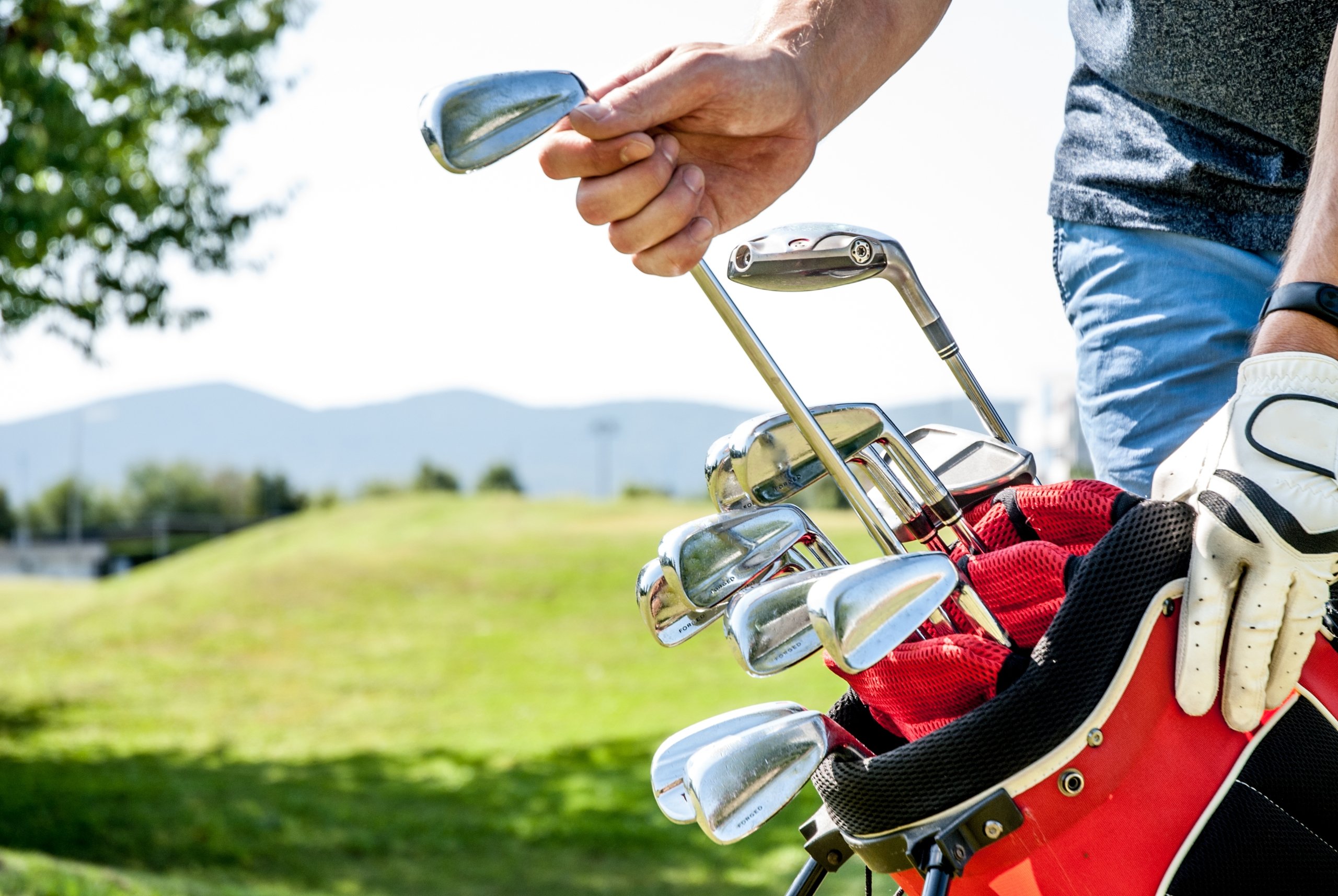 A golfer selects a club from a red golf bag filled with irons on a sunny course.