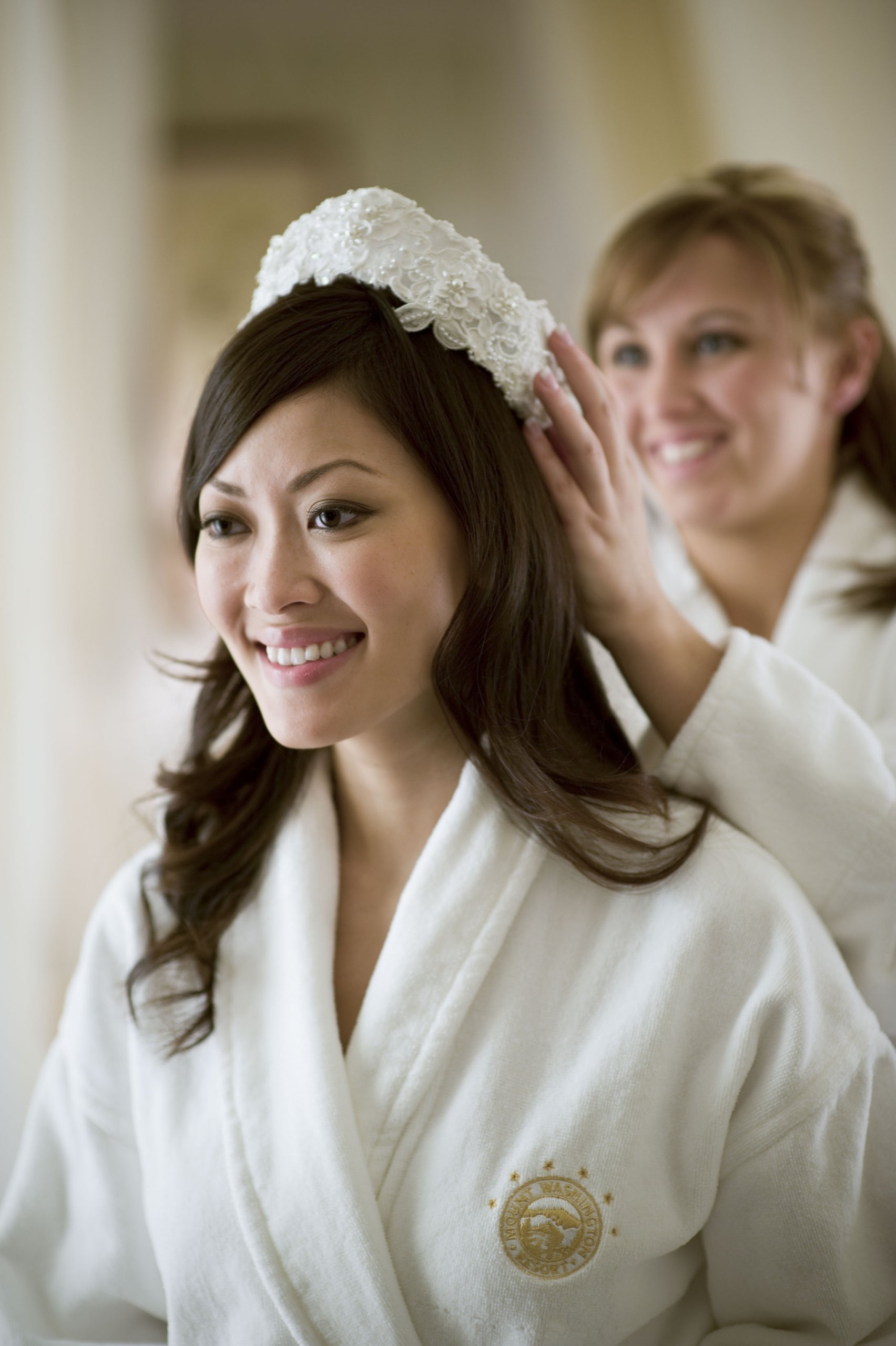 Smiling bride in a white robe has a lace headpiece placed on her hair by a friend in preparation for her wedding at Mount Washington Resort and Spa.