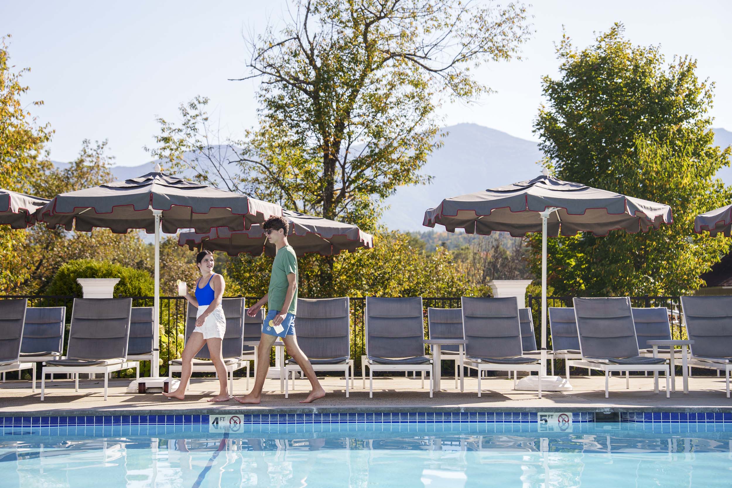 Two people walk barefoot beside a bright blue outdoor pool on a sunny day at Mount Washington Resort and Spa.