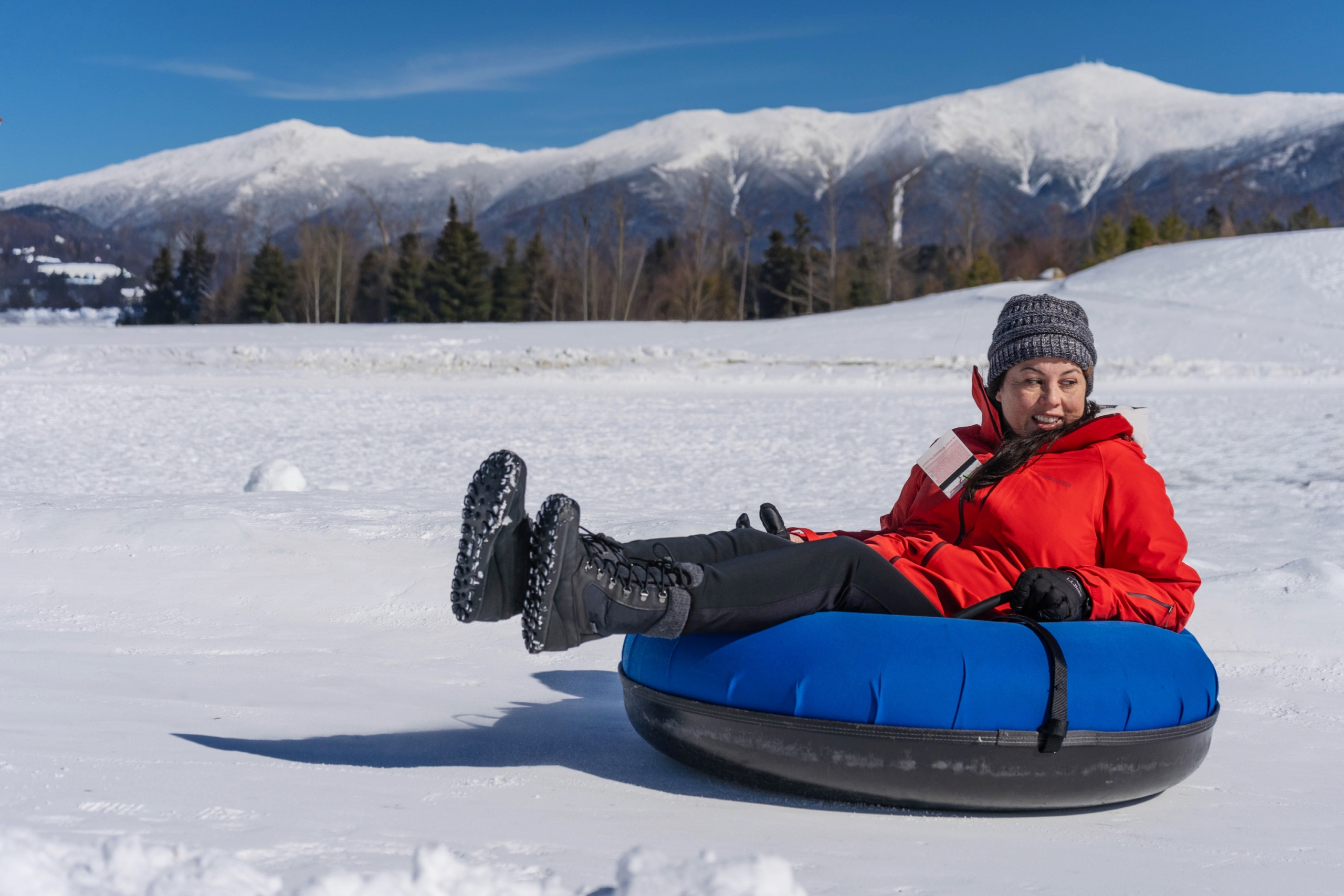 A woman in a red winter jacket smiles while sitting in a snow tube on a snow-covered field at Bretton Woods.