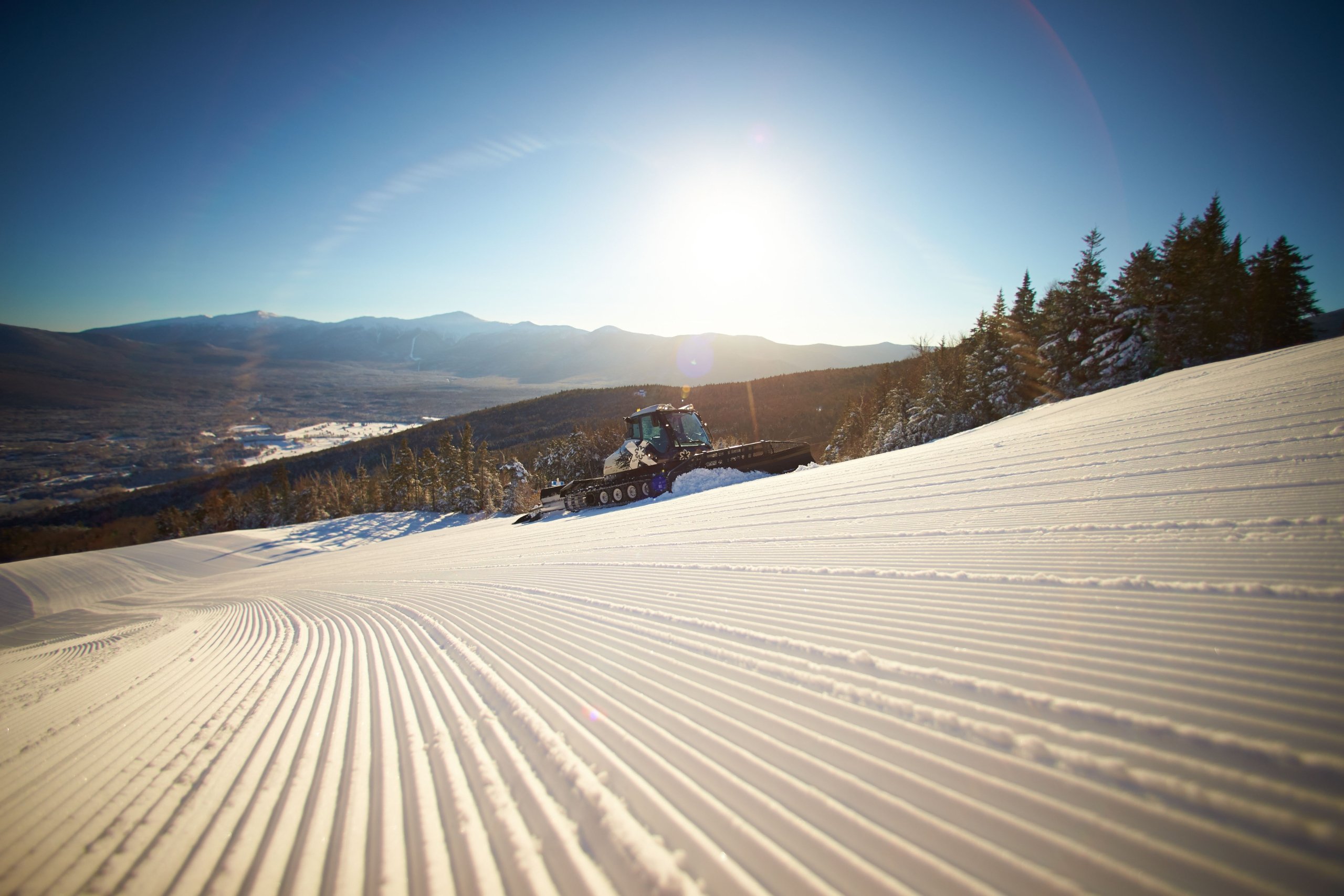 A snow groomer smooths freshly fallen snow on a ski slope under a bright sunrise with mountain views in the distance at Bretton Woods.