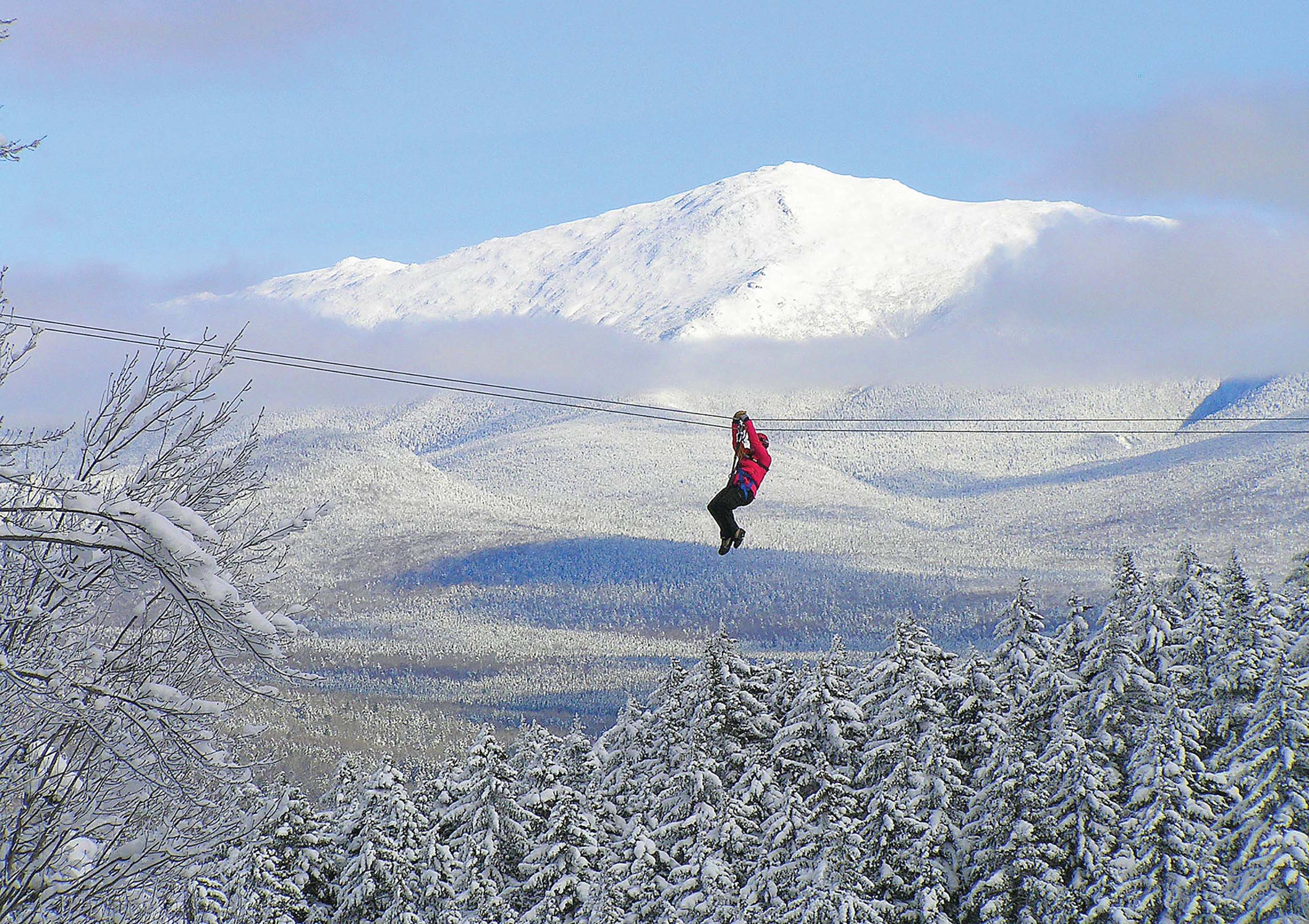 A person hangs by a cable with snow-covered trees beneath them and mountains in the backdrop at Bretton Woods.