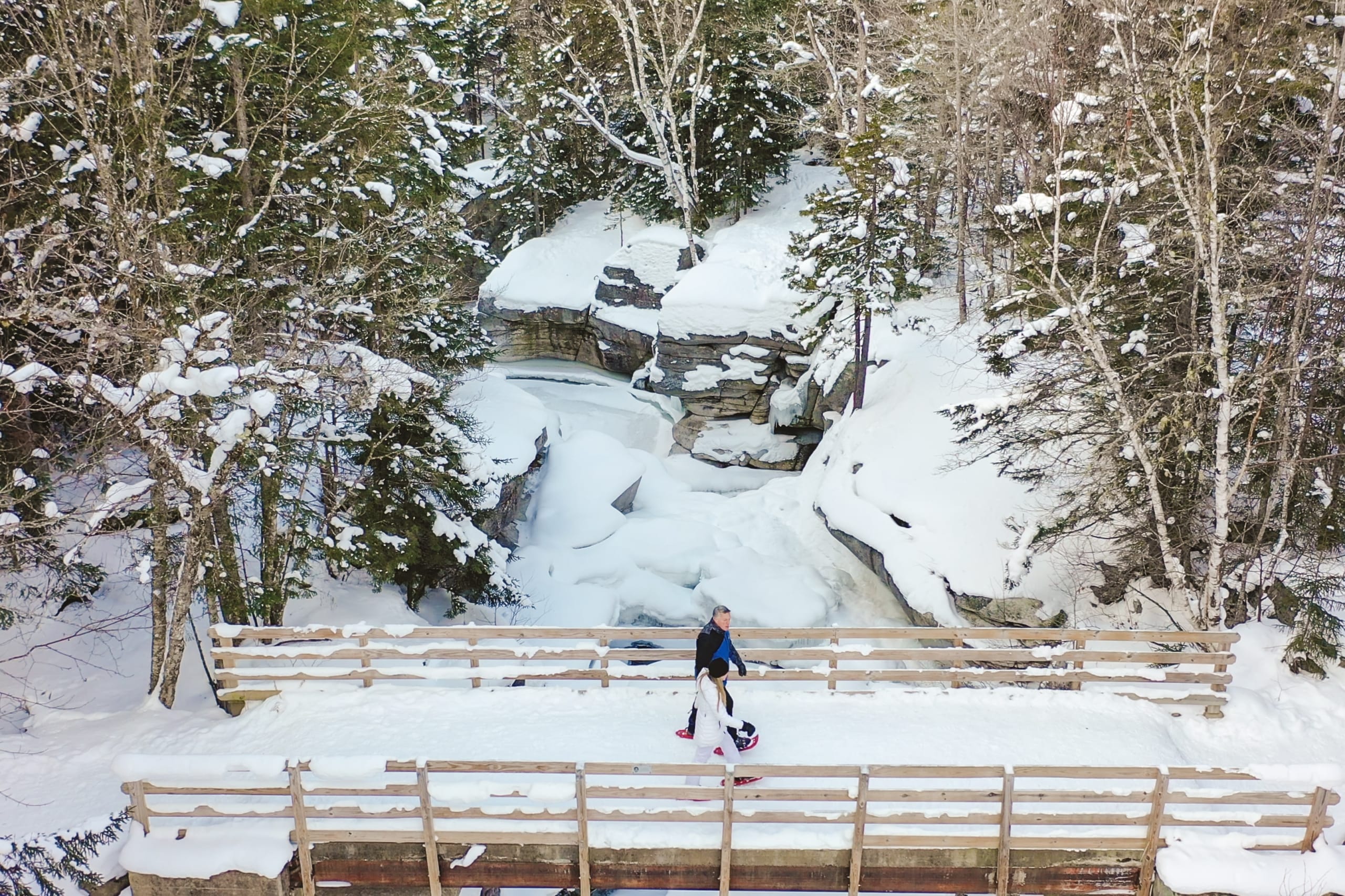 A man and woman walk across a snow-covered bridge in the forest at Mount Washington Resort and Spa.