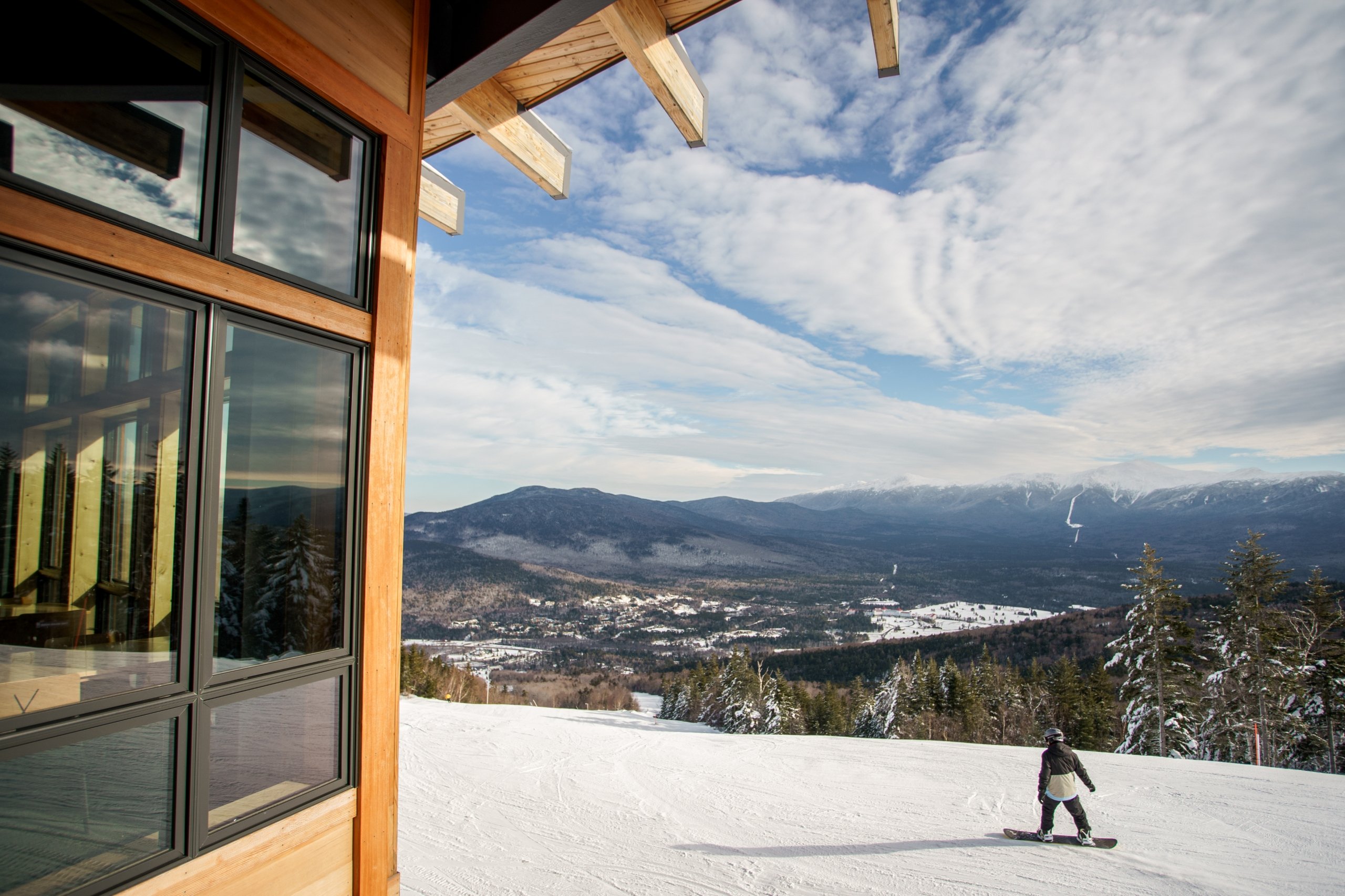 A snowboarder stands on a snowy slope near a wooden lodge, overlooking a scenic mountain valley at Bretton Woods.