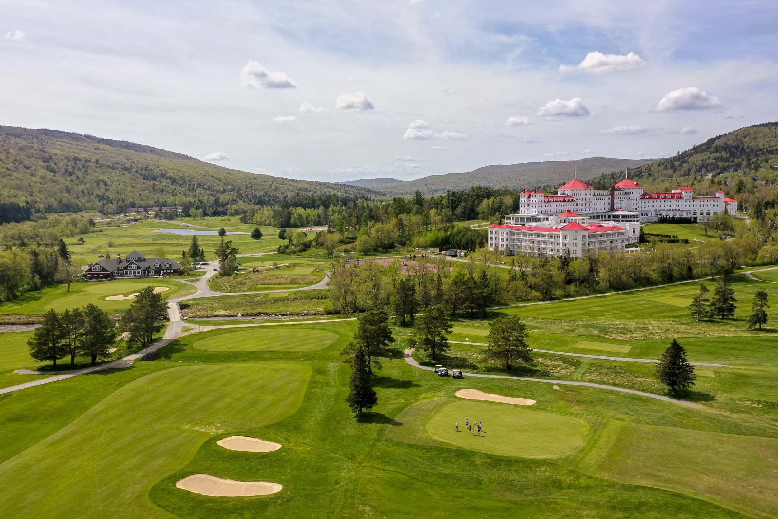 The white exterior and red roofs of Mount Washington Resort and Spa stand out amongst a lush green landscape of mountains and a golf course.