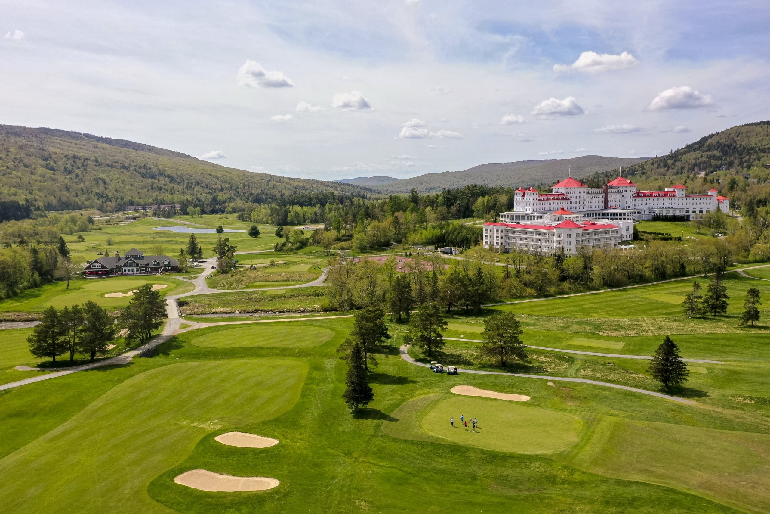 The white exterior and red roofs of Mount Washington Resort and Spa stand out amongst a lush green landscape of mountains and a golf course.
