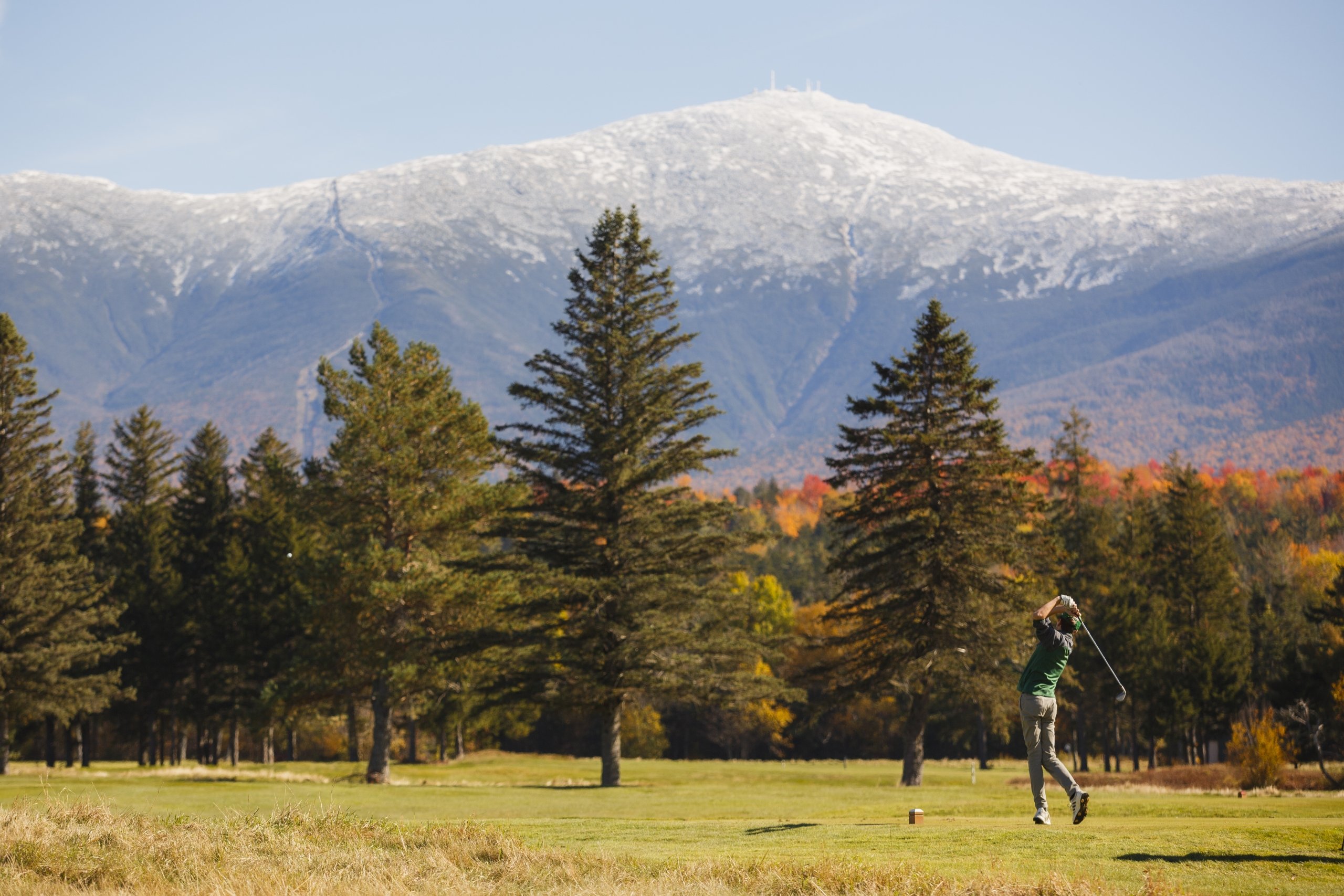A golfer takes a swing at Mount Washington Resort and Spa with a backdrop of colorful autumn trees and a snow-dusted mountain.