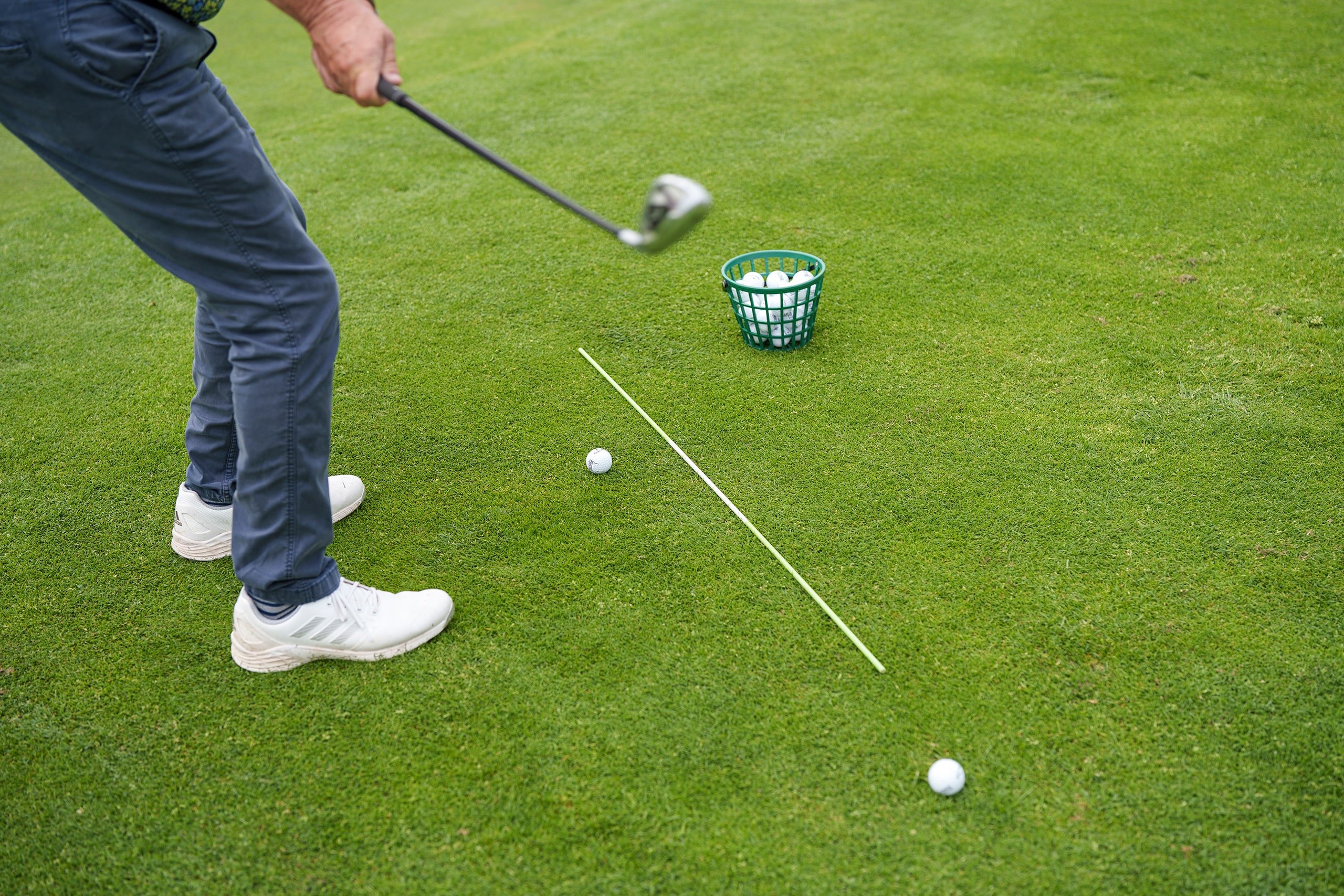A close-up of a golfer positioning themselves to take a swing, with a basket of golf balls at their feet.