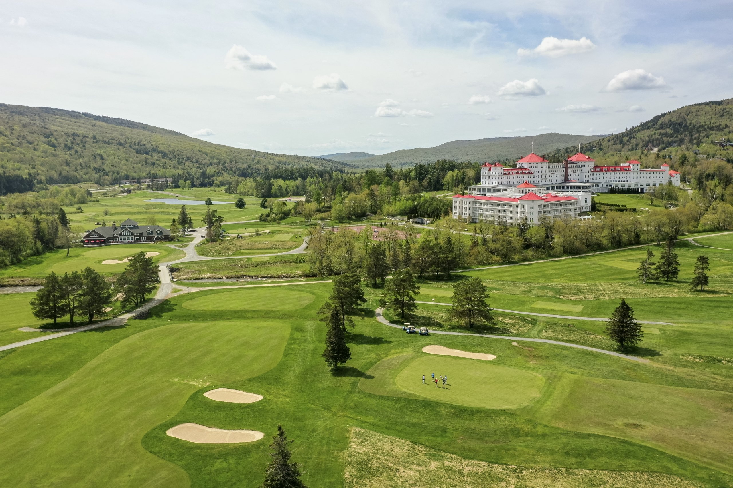 The white exterior and red roofs of Mount Washington Resort and Spa stand out amongst a lush green landscape of mountains and a golf course.
