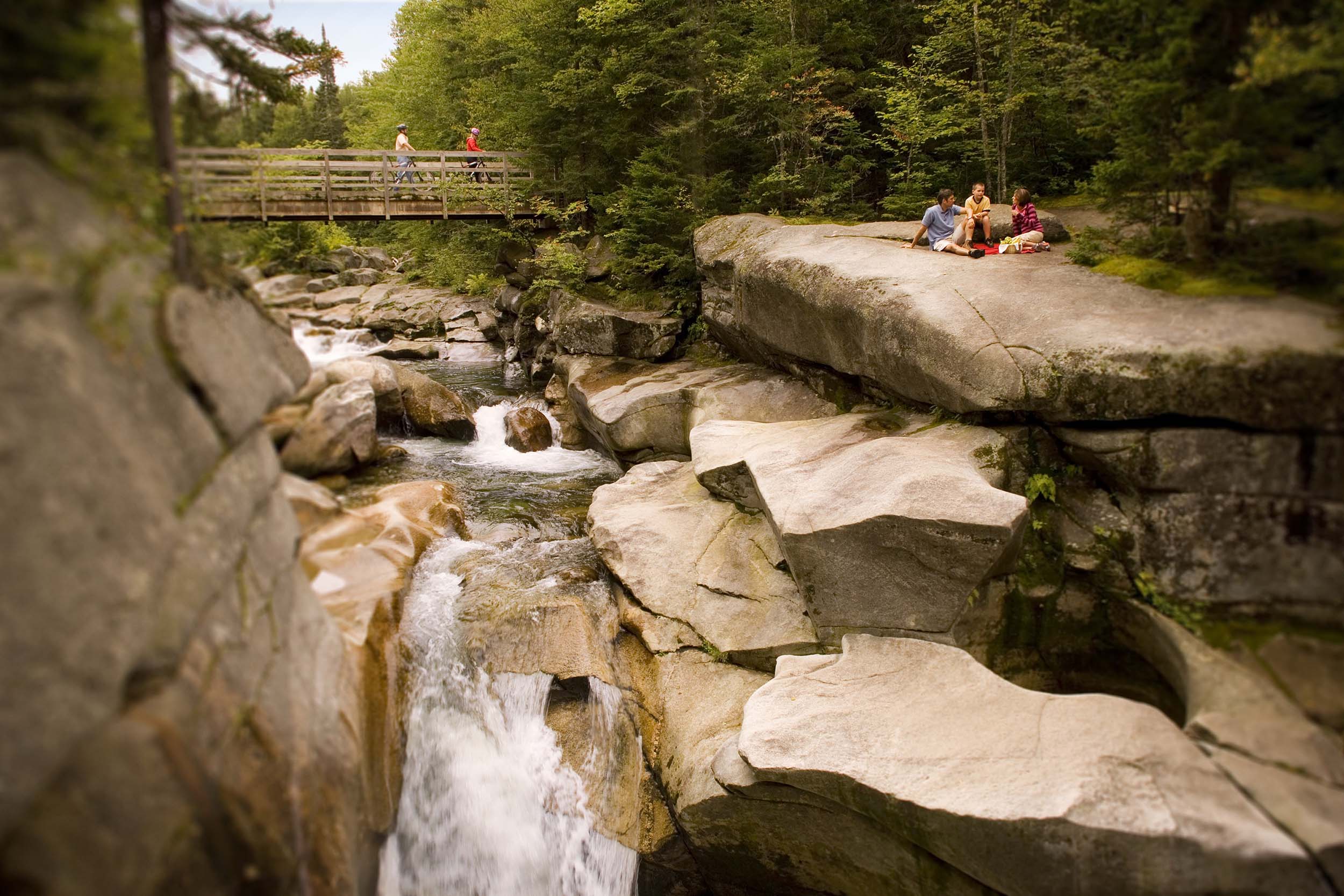 Hikers relax on rocks while other hikers pass over the bridge on the Summer trails at Bretton Woods.