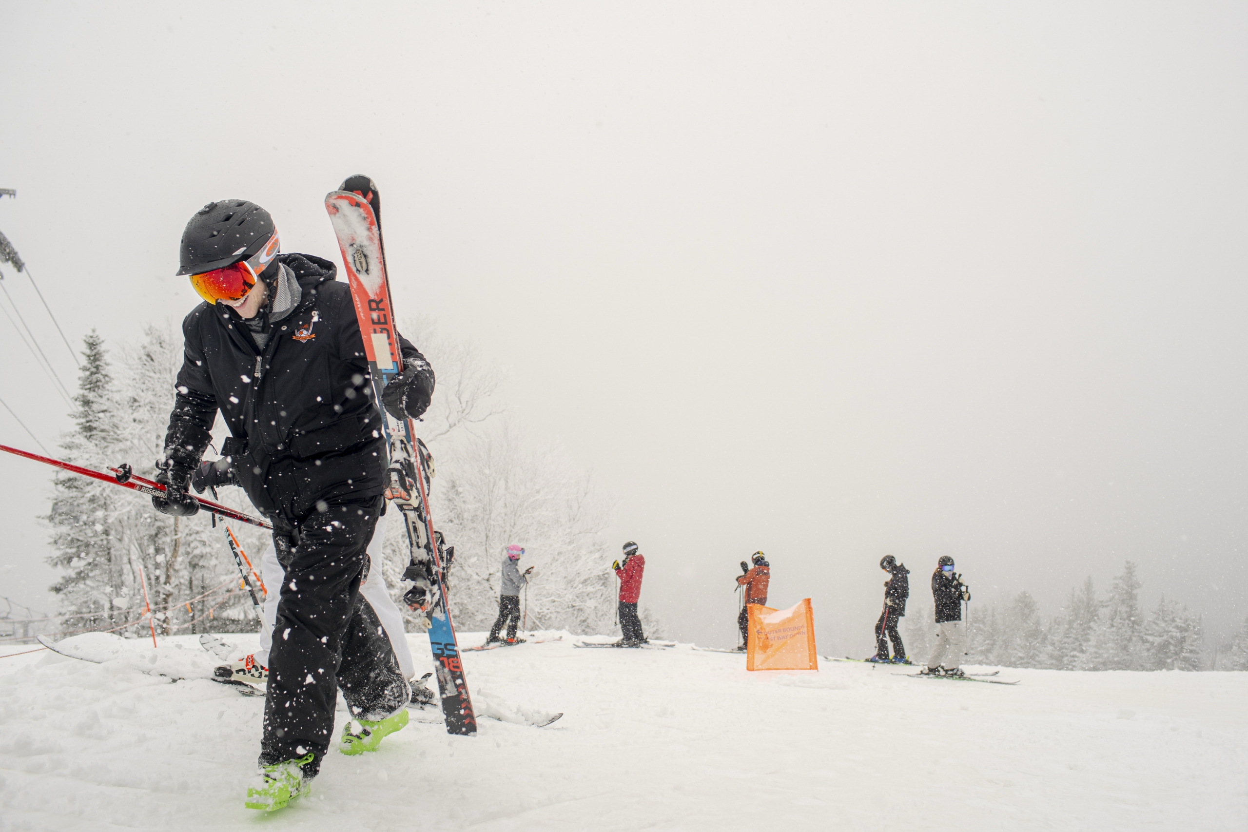 A skier carries their skis while a group of skiers gathers in the snowy background at Bretton Woods.
