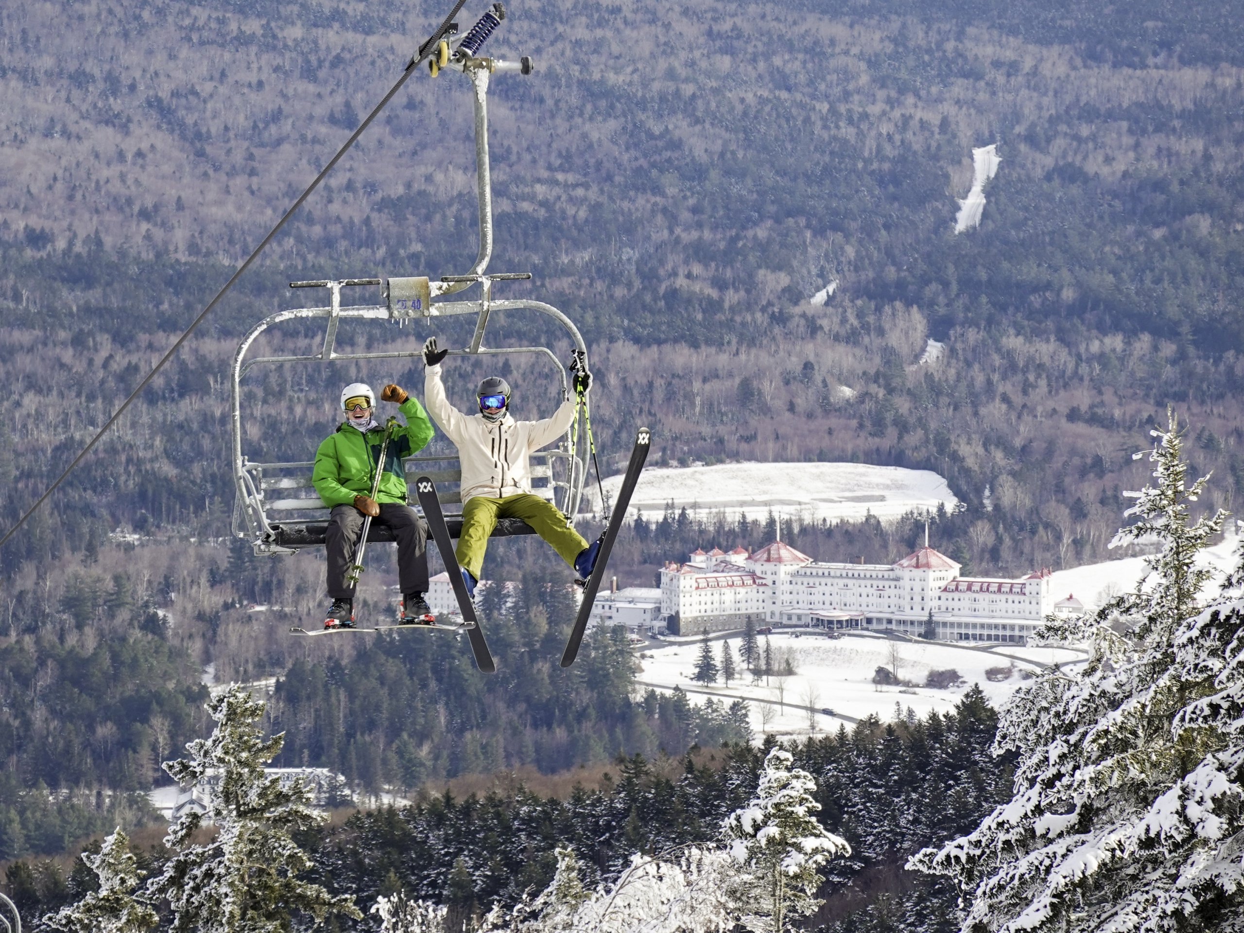 Two skiers riding a chairlift high above a scenic valley at Bretton Woods, with Mount Washington Resort & Spa visible against the backdrop of snow-dusted hills.