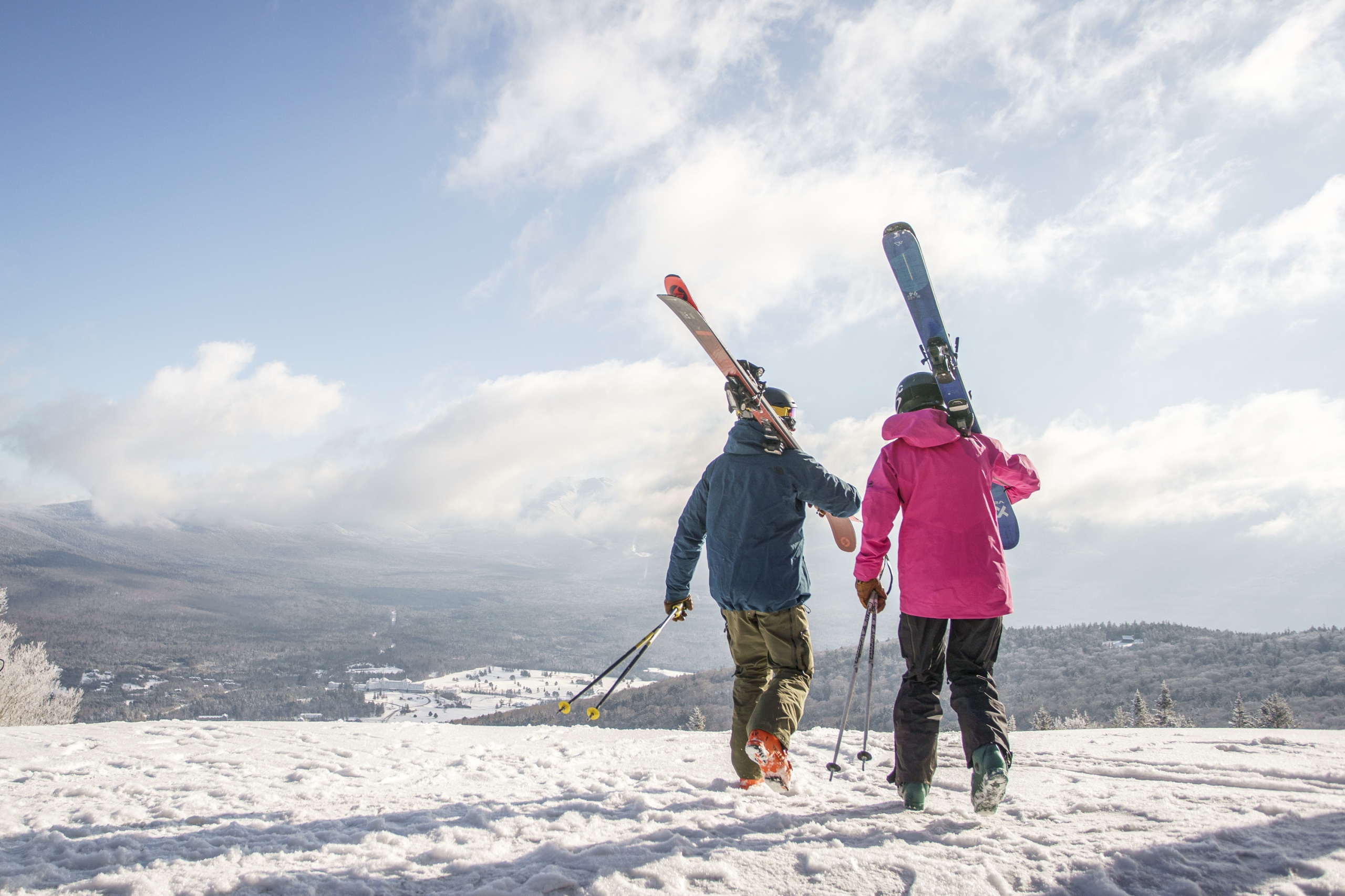 Two skiers walk across a snowy mountain ridge at Bretton Woods, carrying their skis on their shoulders.