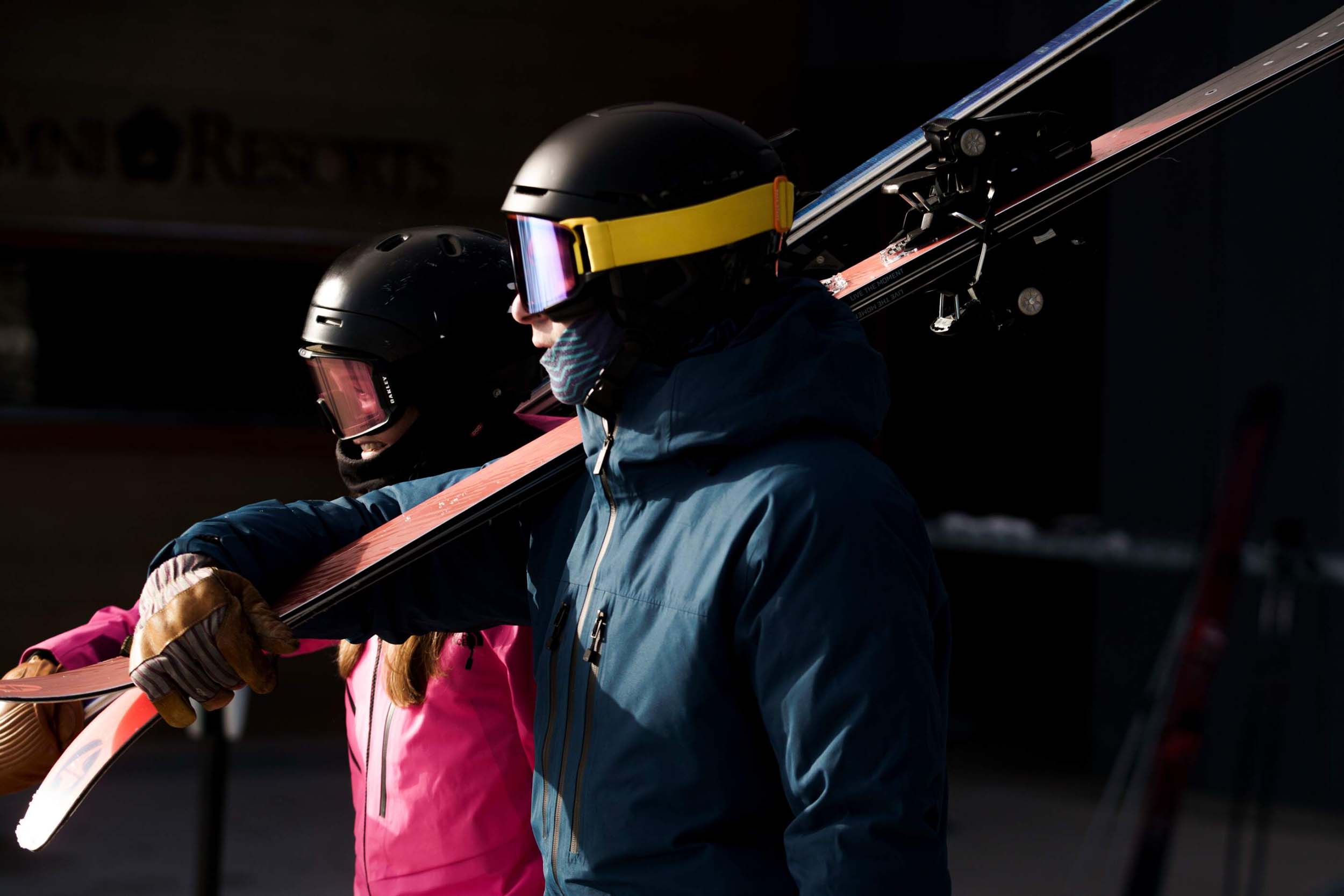 Two skiers wearing goggles and helmets carry their skis over their shoulders.