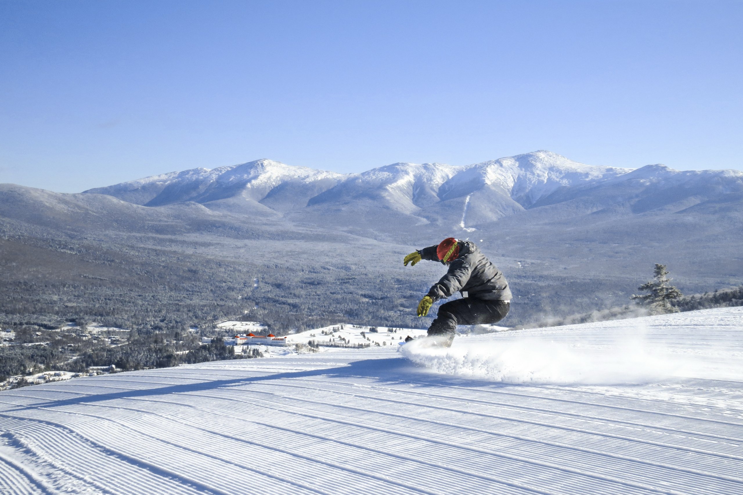 A snowboarder carves through fresh snow on a sunny day at Bretton Woods, with snow-covered mountains in the distance.