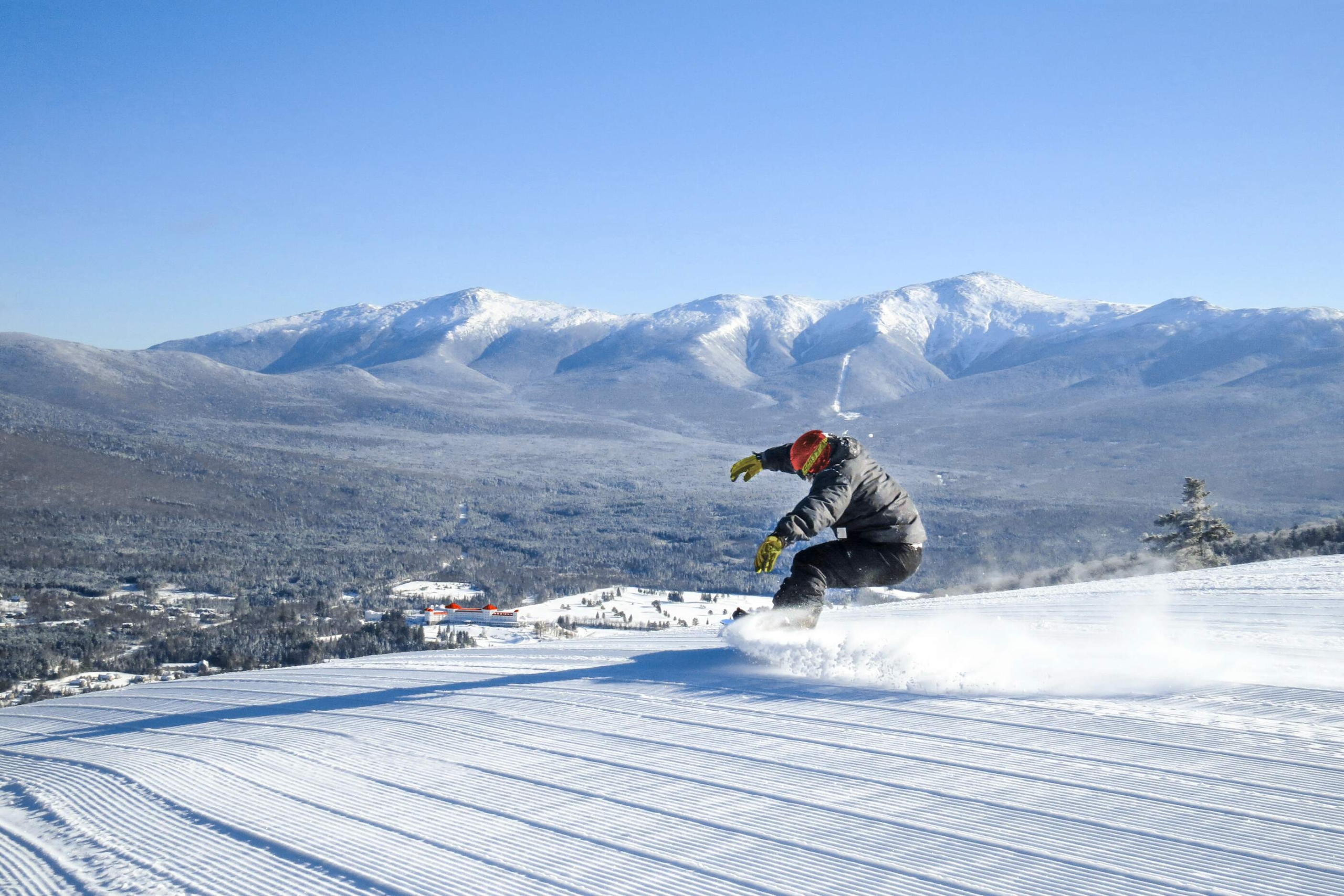 A snowboarder carves through fresh snow on a sunny day at Bretton Woods, with snow-covered mountains in the distance.