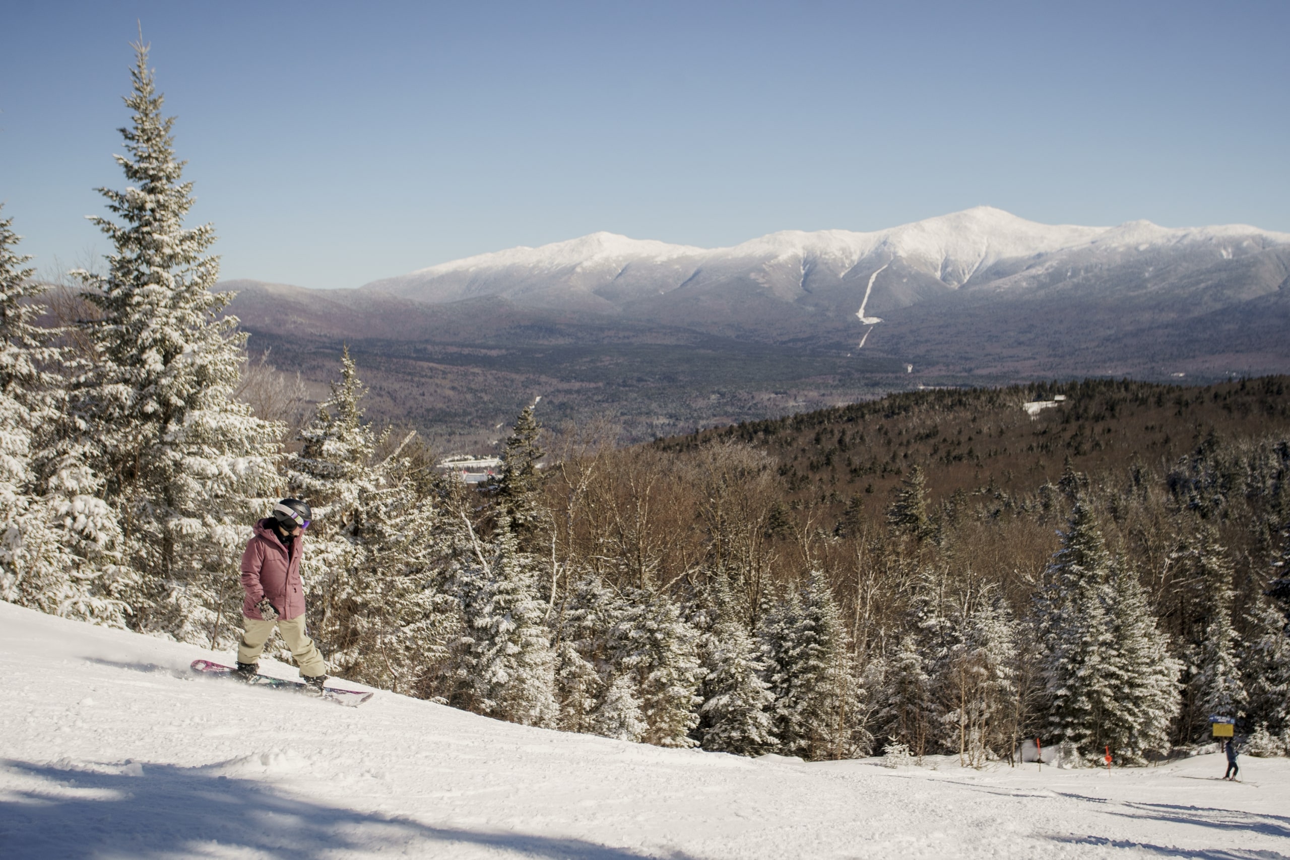 A snowboarder descends a hill with a lush forest and snow-covered mountains in the background at Bretton Woods.