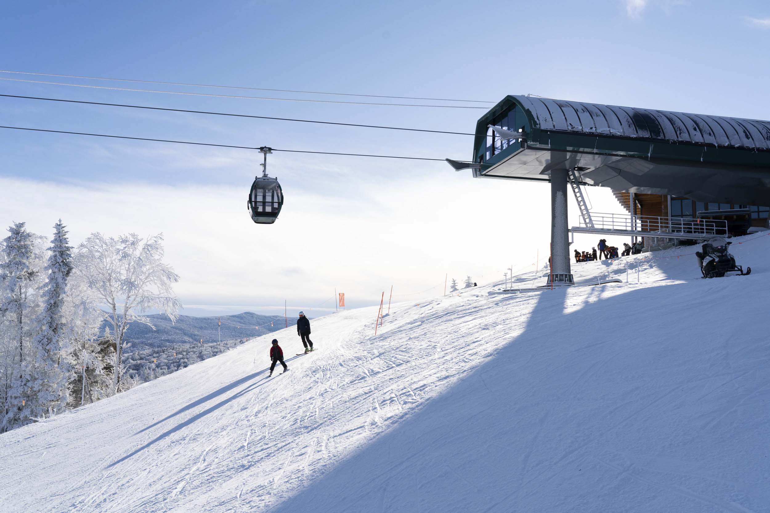 Skiers make their way down a snowy slope near a gondola lift station under a bright blue sky at Bretton Woods.