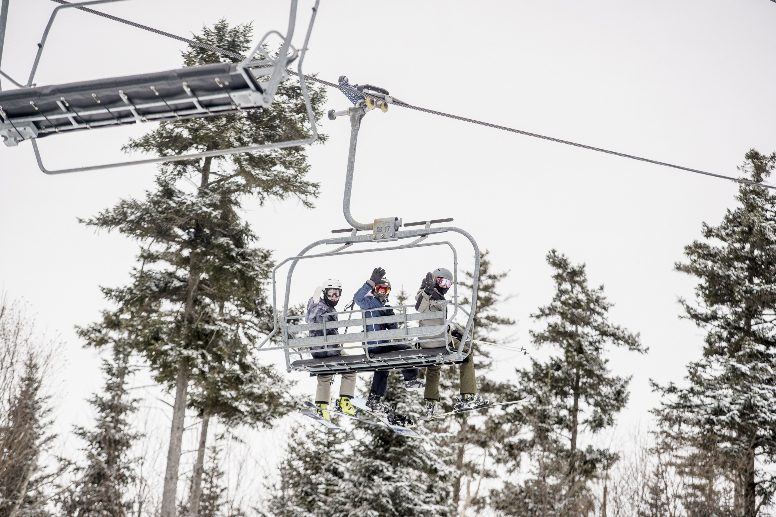 Three skiers ride a chairlift at Bretton Woods above snow-covered trees, waving toward the camera.