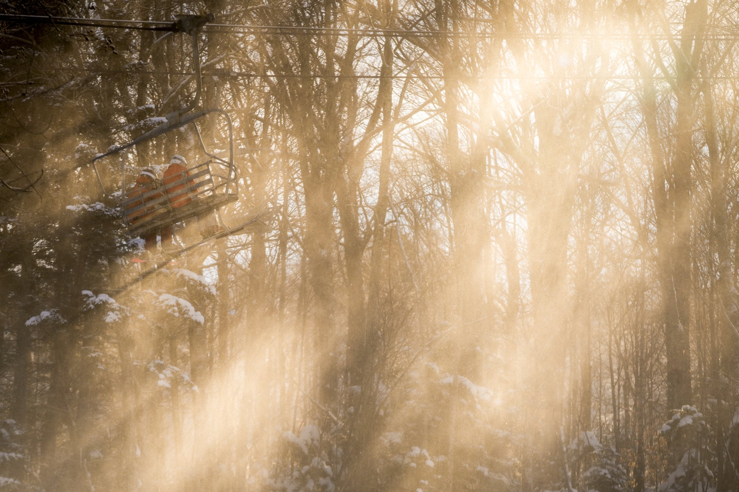 Two skiers ride a chairlift through a forest as golden sunlight streams through the trees at Bretton Woods.