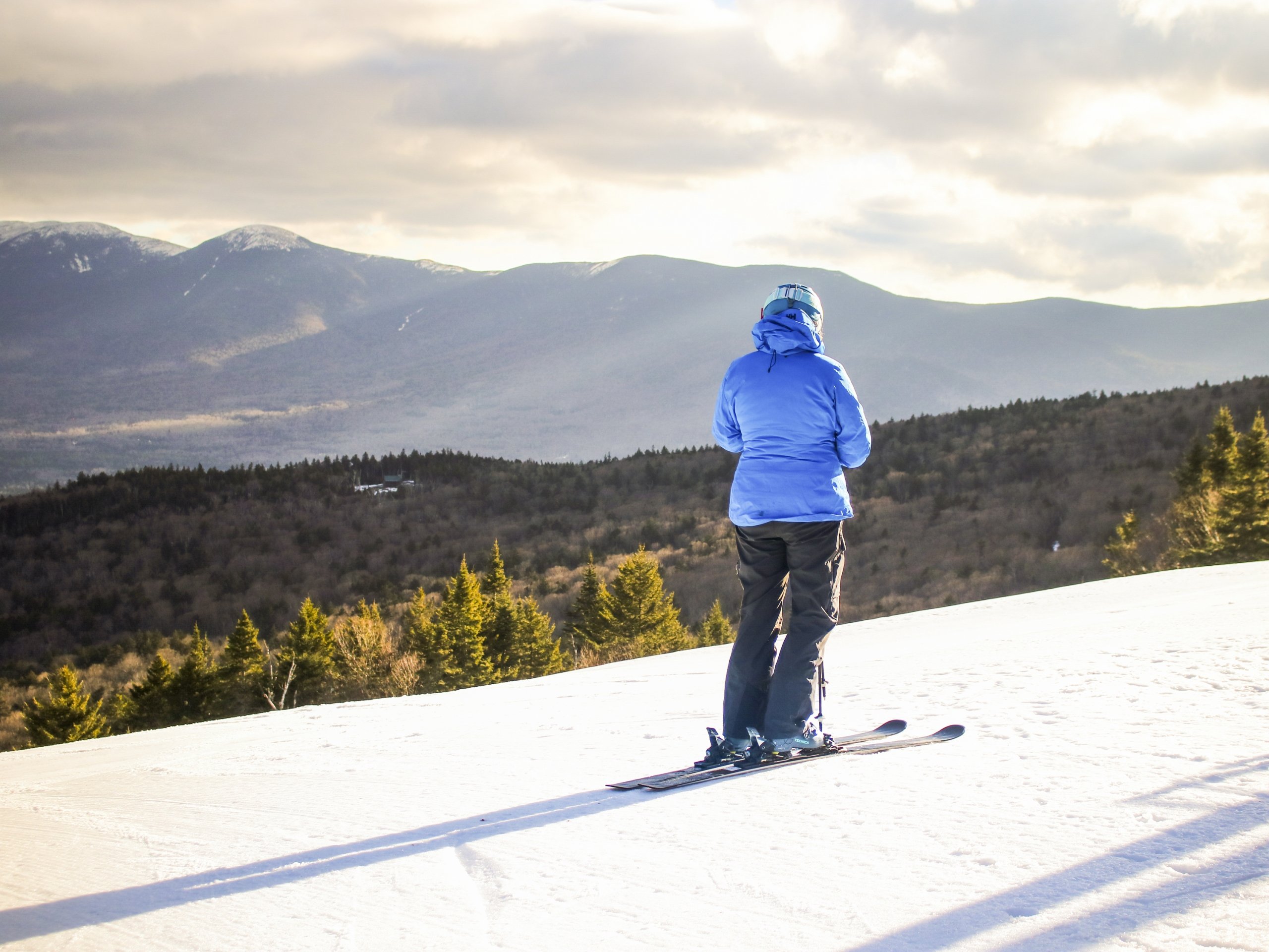 A skier stands on a snowy slope overlooking a valley of evergreen trees and distant mountains at Bretton Woods.