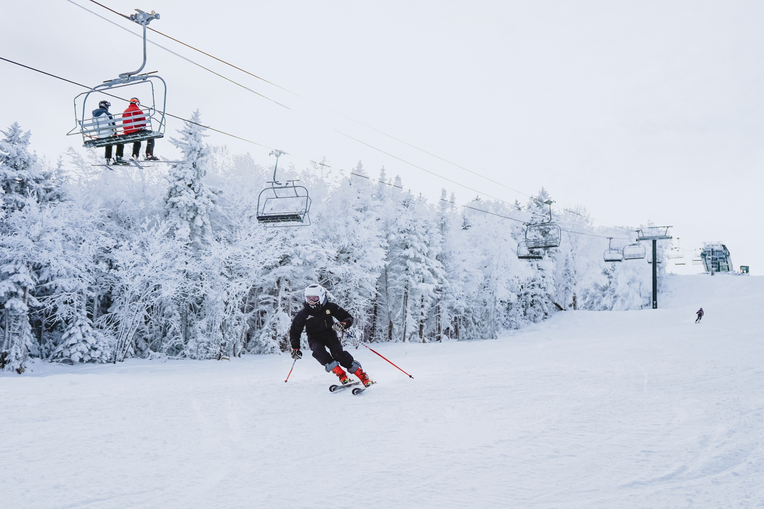 A skier racing down a snowy ski hill while other skiers ride the chairlift above at Bretton Woods.