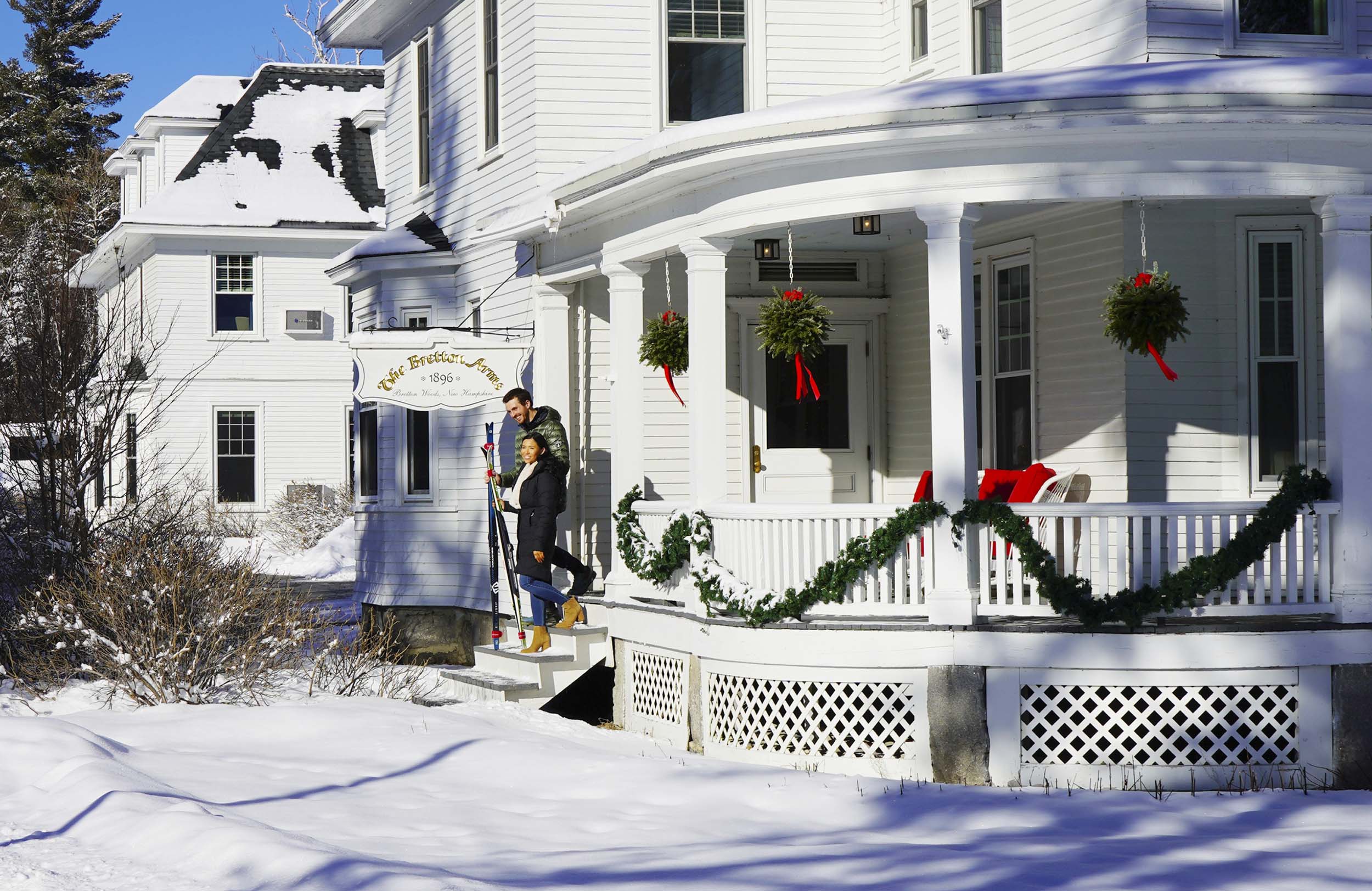 A man and woman carrying cross-country skis walk down the snowy steps of The Bretton Woods Inn with its porch decorated with evergreen garland and hanging wreaths with red ribbons.