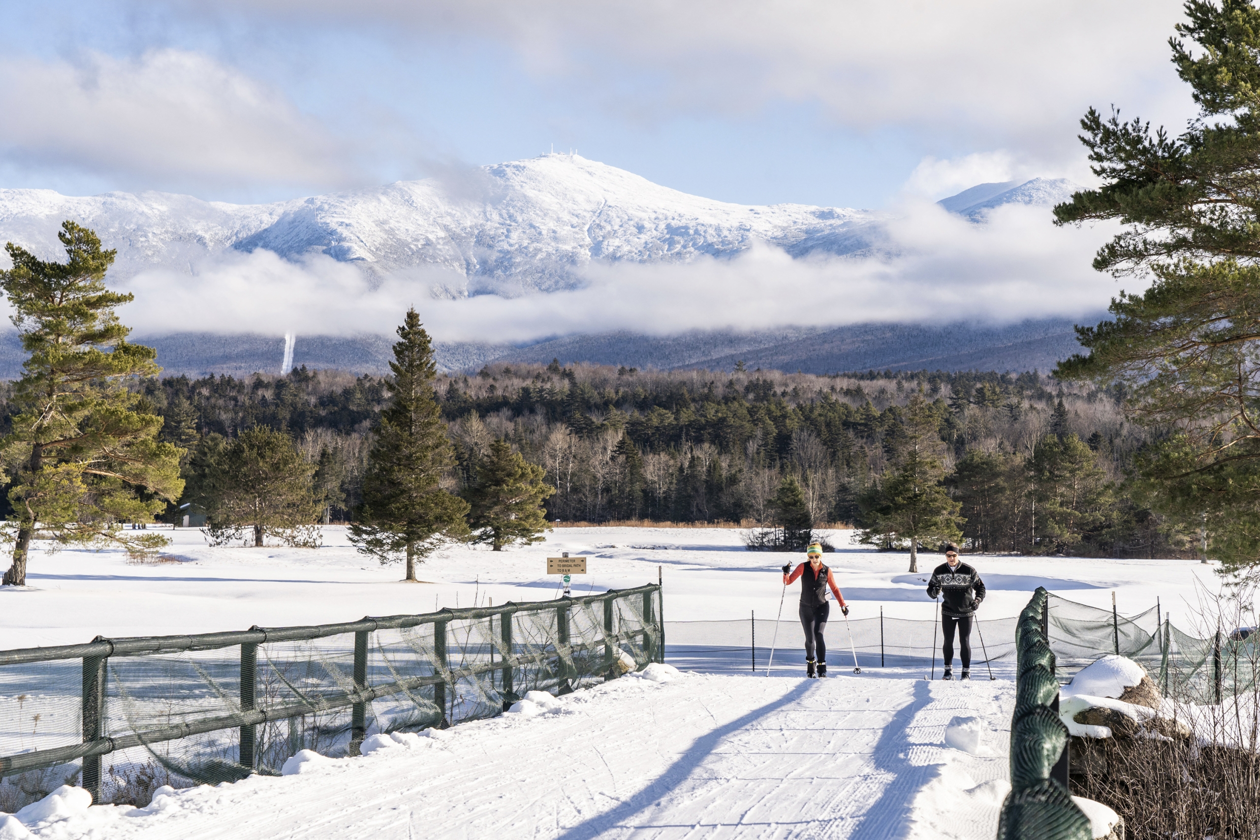 Two people cross-country ski on a groomed track over a bridge at Bretton Woods, with snow-capped mountains in the distance.