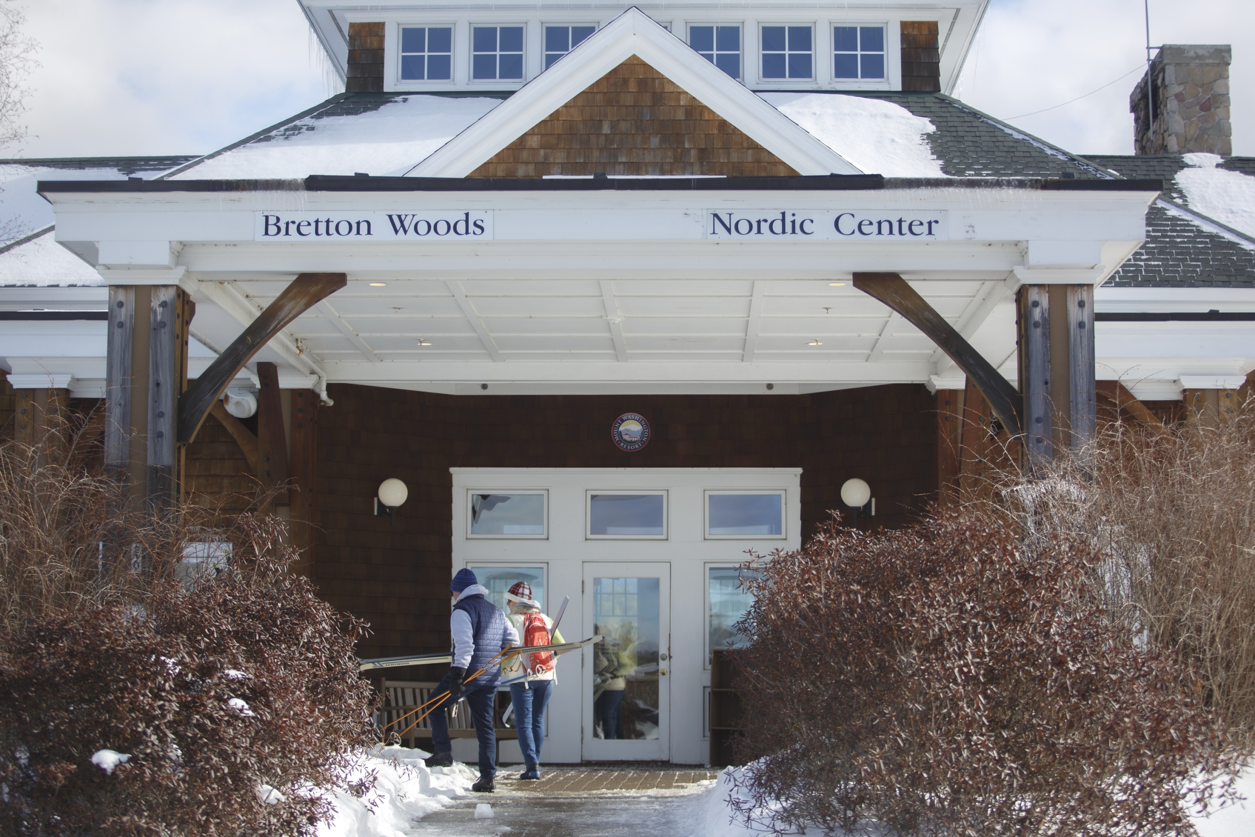 Two people carrying skis are walking up a snowy path toward the entrance of Bretton Woods Nordic Center.