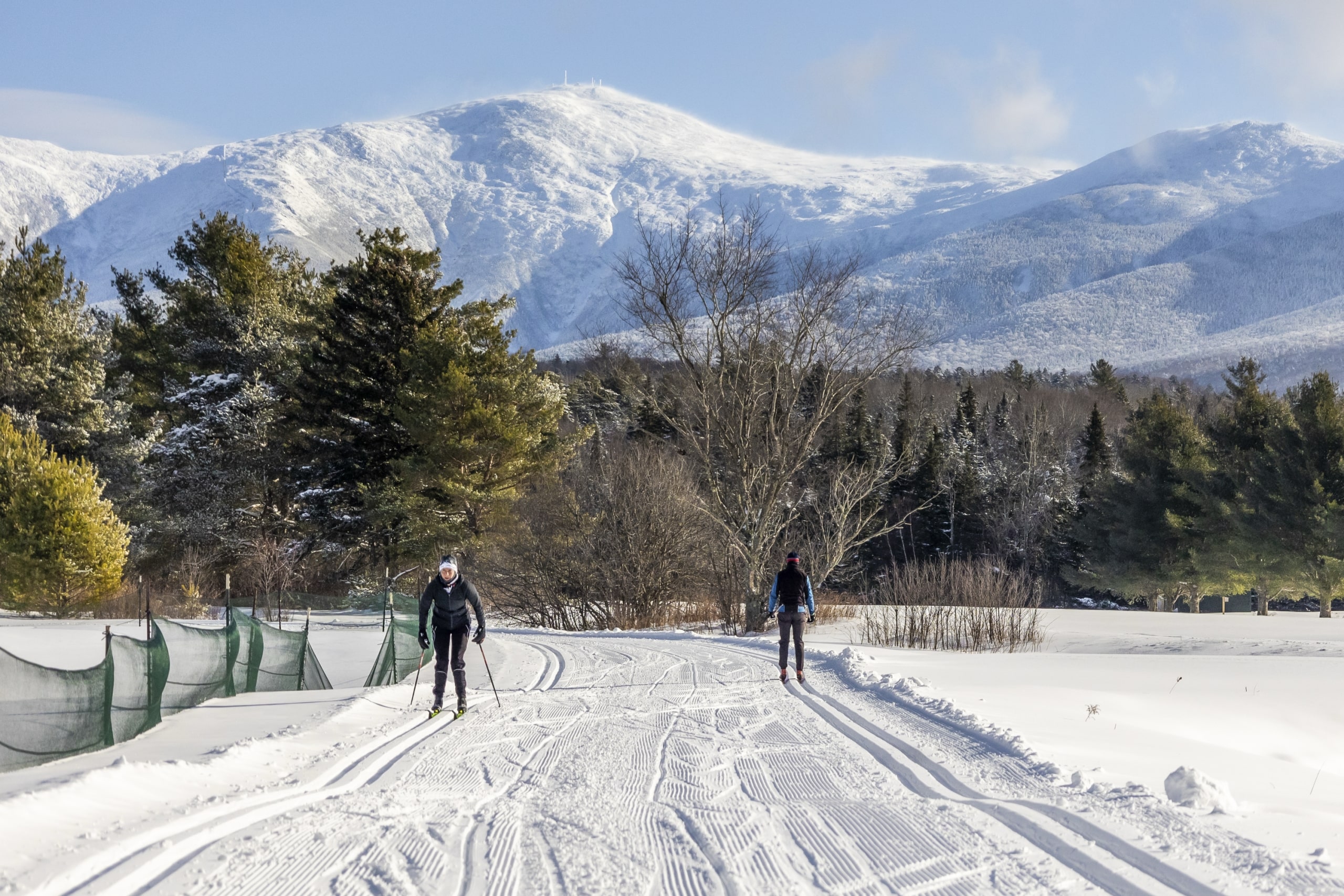 Two people cross-country skiing on a wide, groomed trail at Bretton Woods with snow-covered mountains in the background.