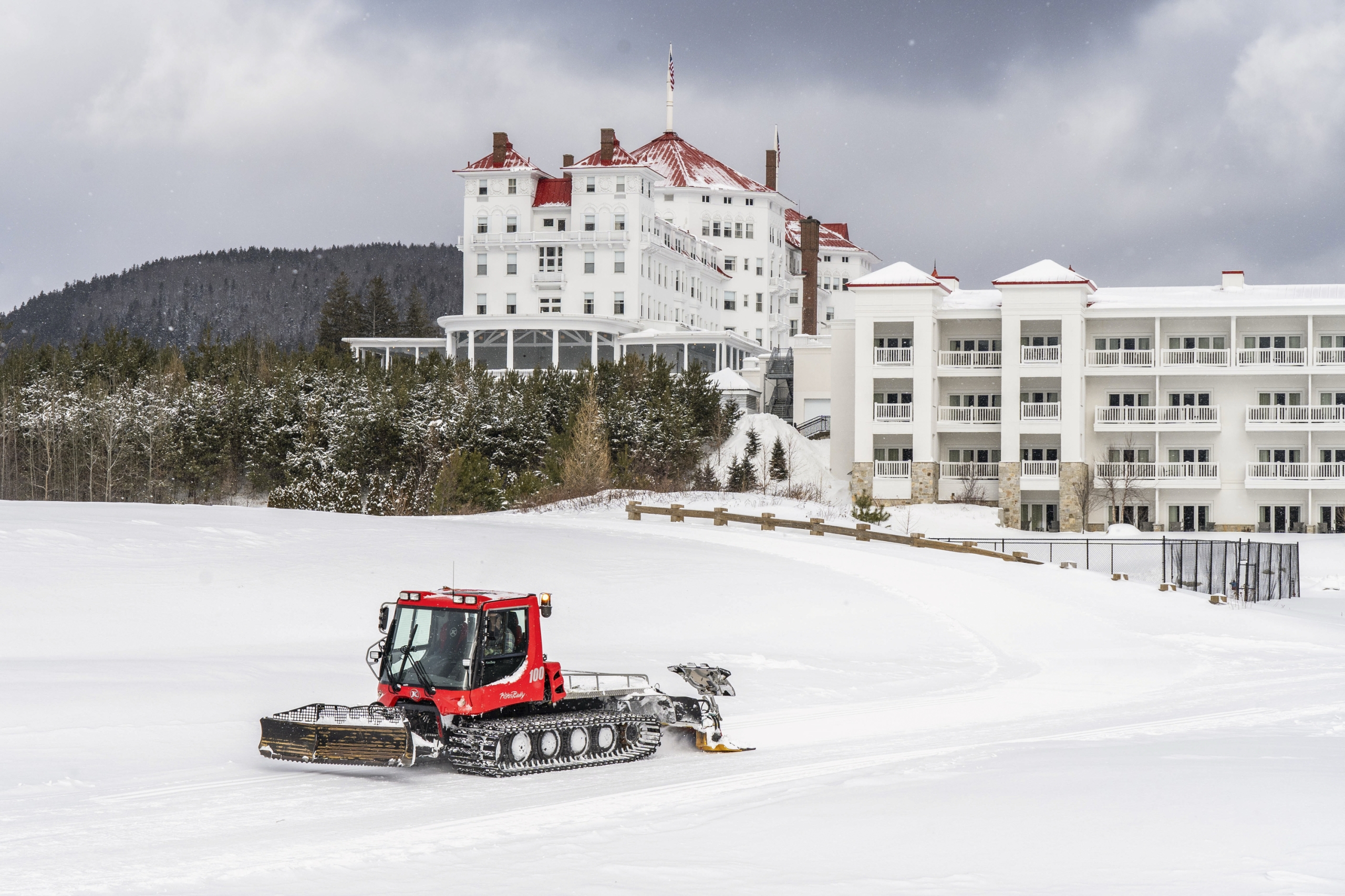 A red snow groomer grooming a snow-covered field with the iconic Mount Washington Resor and Spa standing prominently against a cloudy sky.