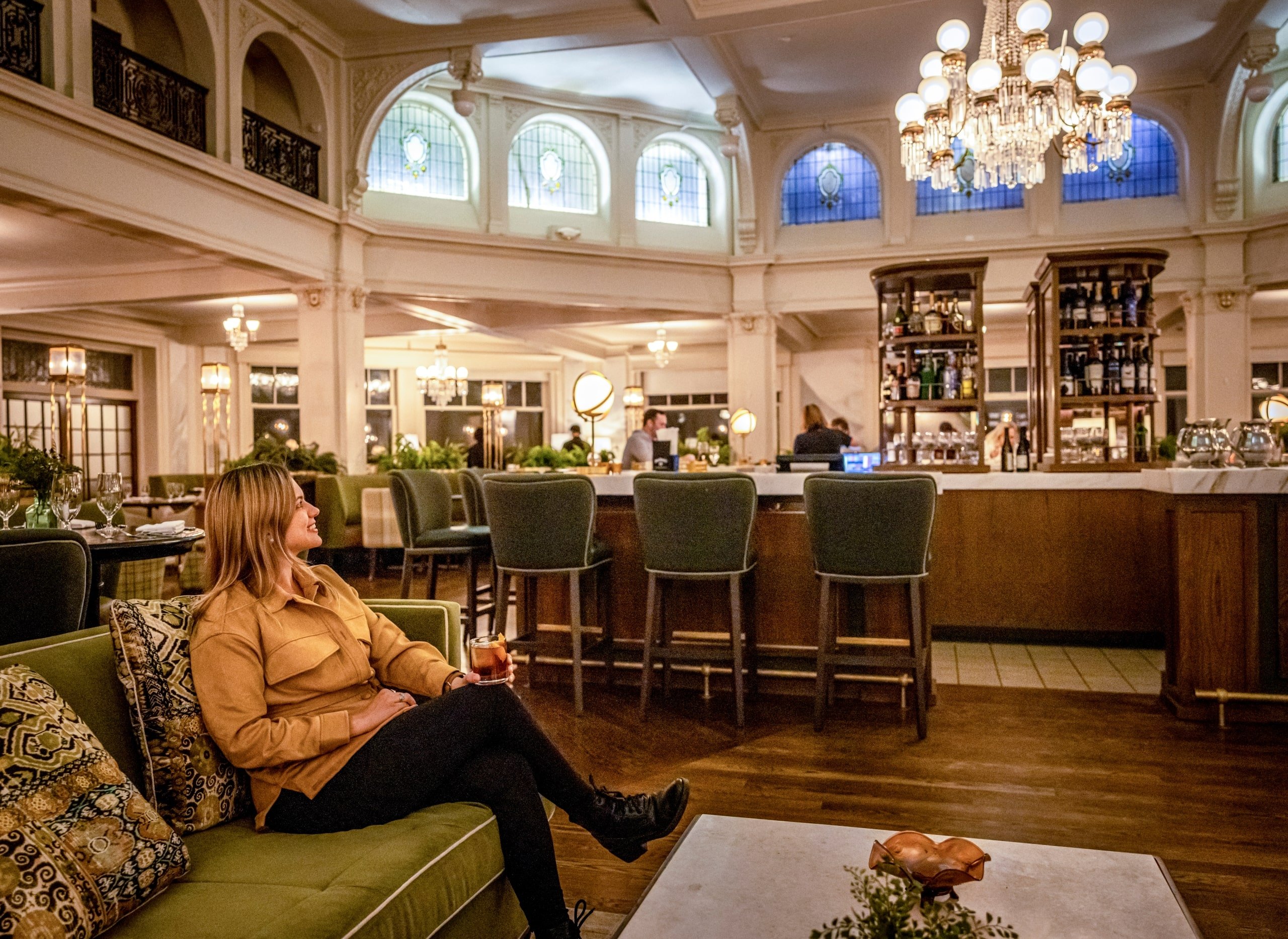 A woman relaxing with a drink in the elegant hotel bar with chandeliers and classic décor at Mount Washington Resort and Spa.