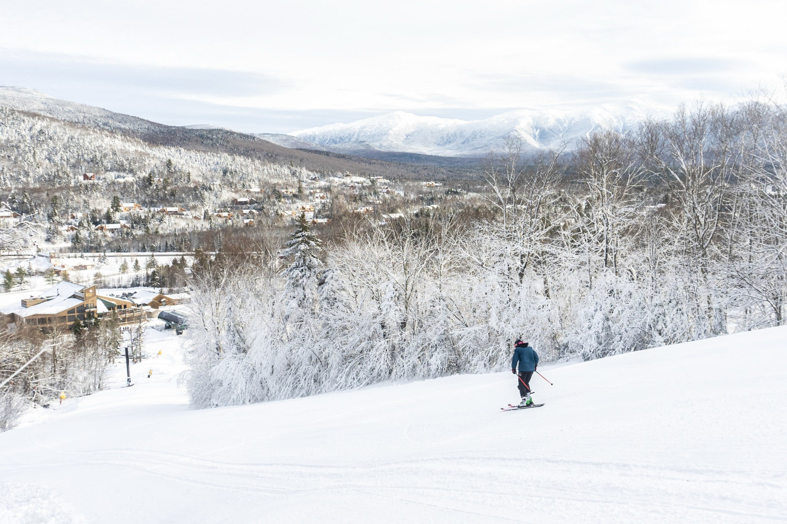 A skier glides down the snowy trail at Bretton Woods with a view of the ski lodge and snow-covered mountains in the distance.