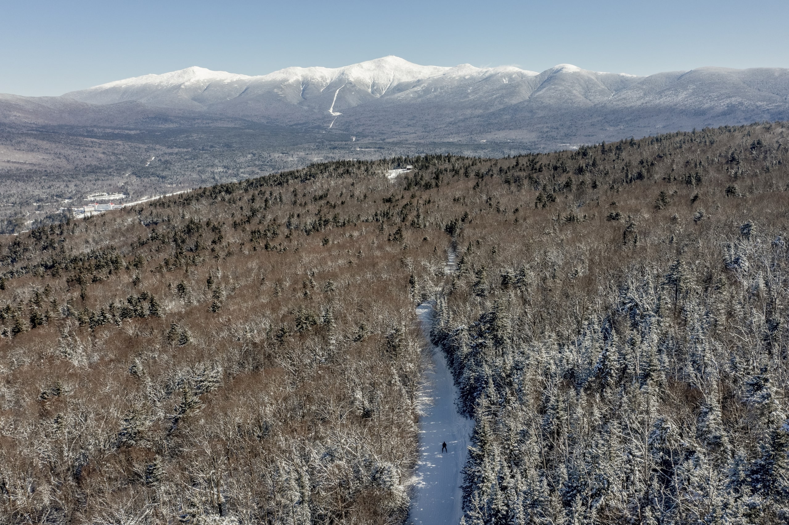 An aerial view of a snow-covered forest with a narrow ski trail cutting through the trees, leading toward a backdrop of snow-capped mountains at Bretton Woods.
