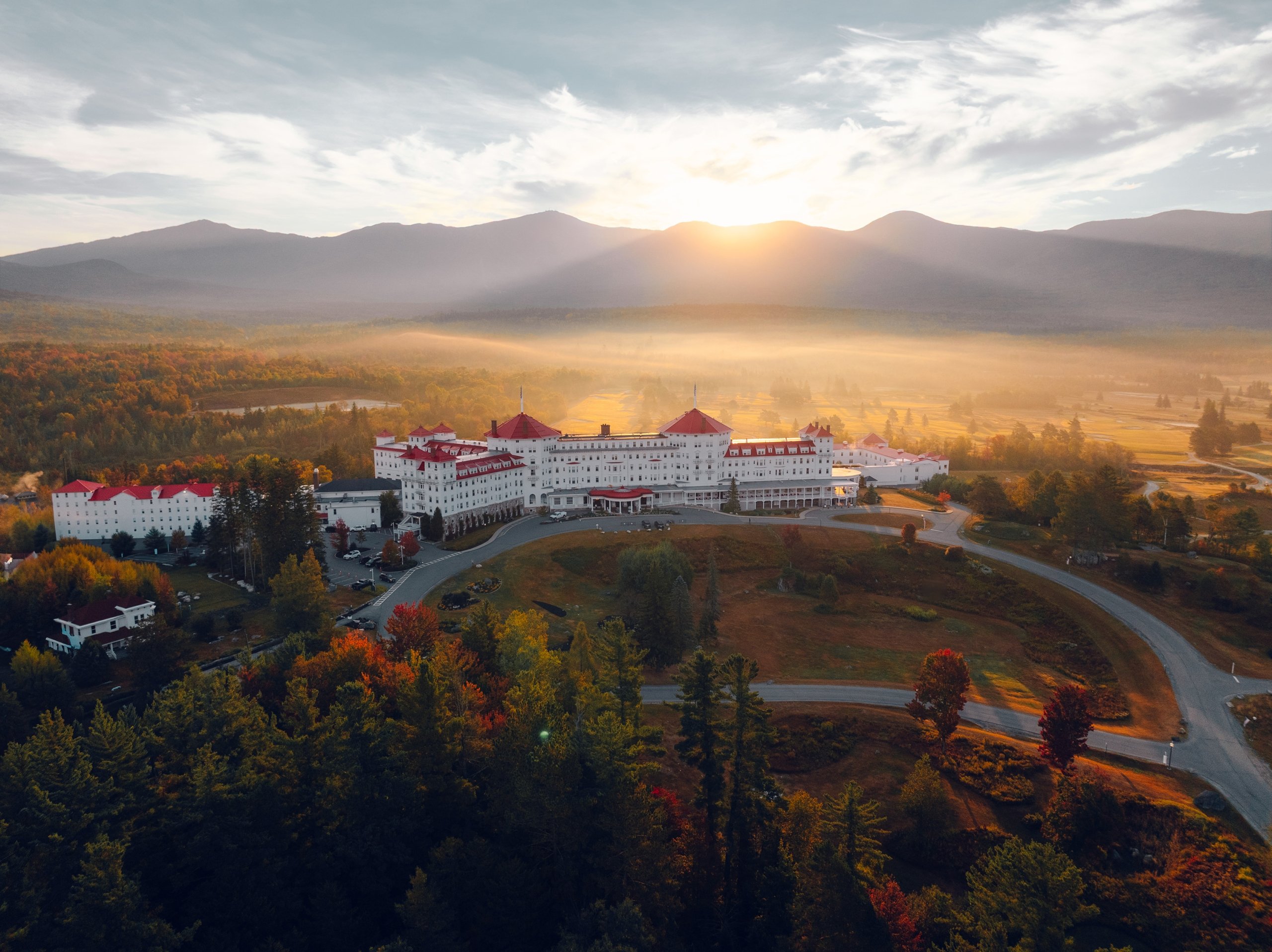 Omni Mount Washington Resort and Spa stands amid fall foliage at sunrise with Mount Washington in the distance and fog on the golf course.